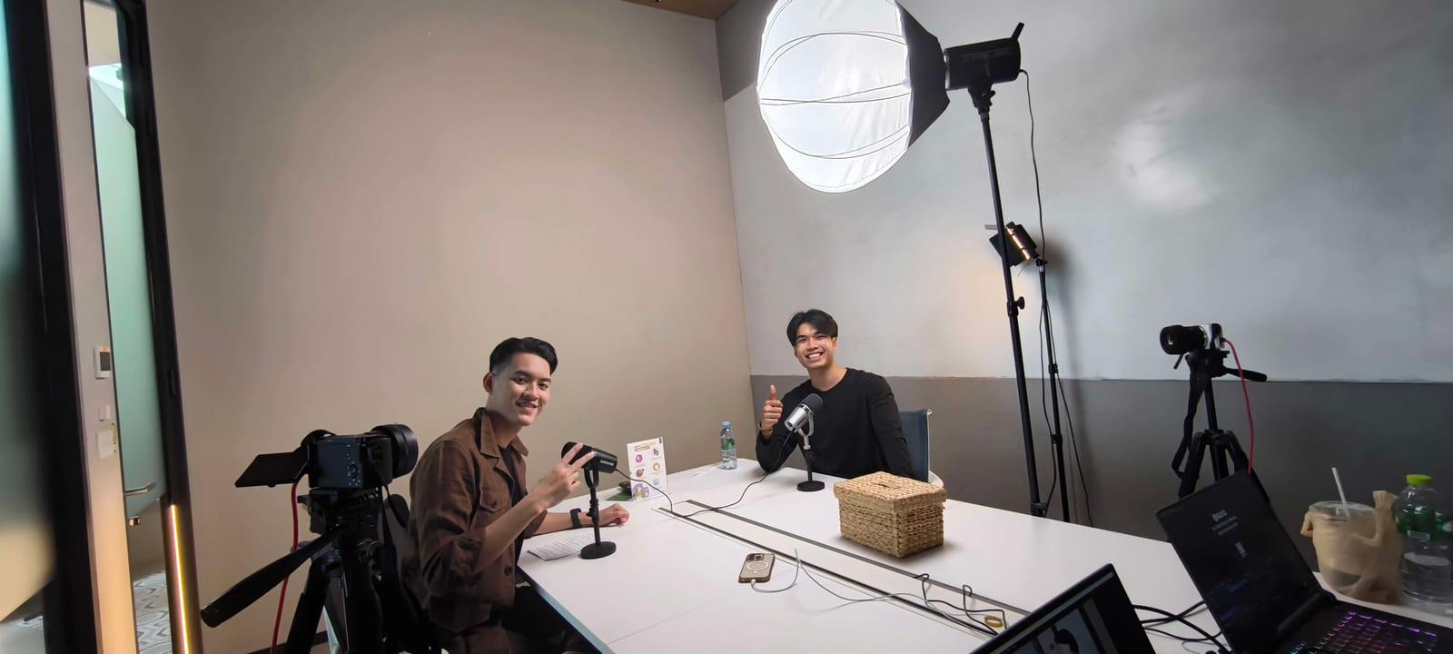 ผมกับแขกรับเชิญนั่งคุยกันหน้าไมโครโฟน ใต้ไฟสตูดิโอ โดยมีกล้องตั้งถ่ายอยู่ด้านข้าง