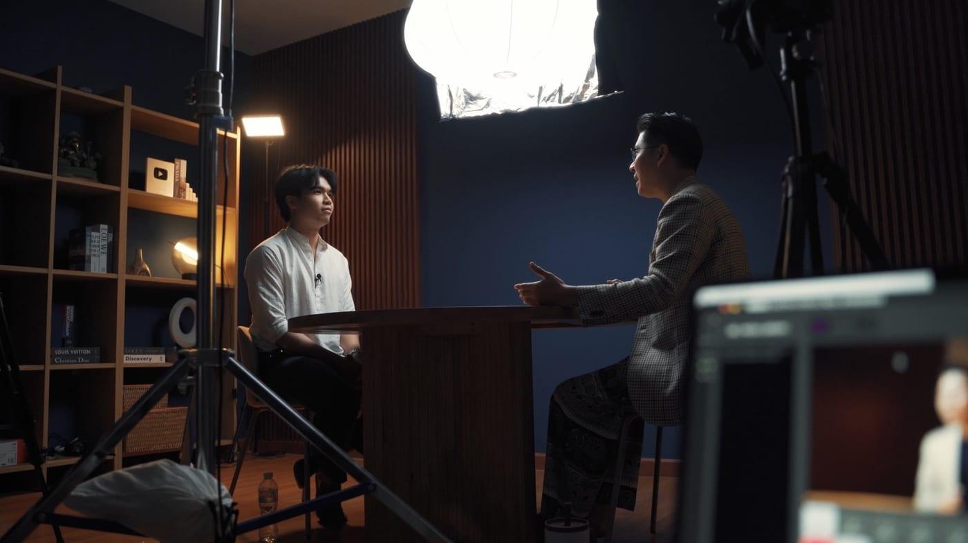 มุมกว้างของกองถ่าย ผู้ร่วมรายการสองคนนั่งคุยกันที่โต๊ะ ล้อมด้วยไฟและกล้องของทีมงาน